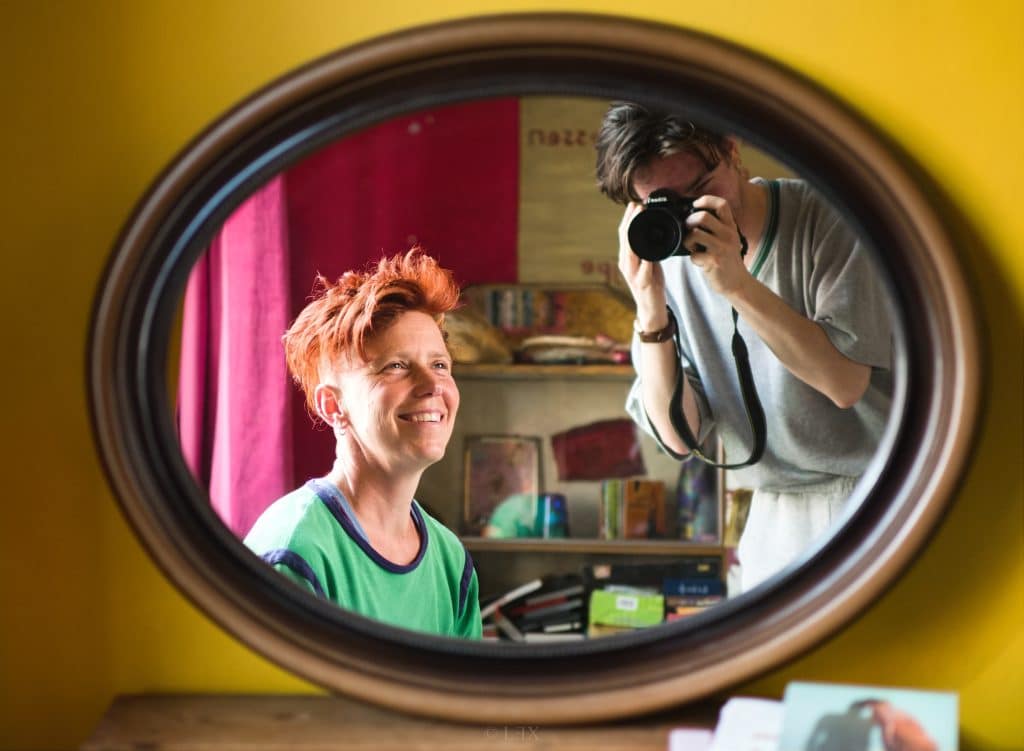 Mees is te zien via een ovale spiegel. Naast hen is in de achtergrond Lex, de fotograaf, te zien met de camera. Mees kijkt naar Lex en lacht.